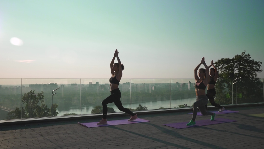 Female silhouettes stand in yoga virabhadrasana warrior pose at group class outdoor on sunrise. Fit healthy young women do yoga fitness exercise meditating together at retreat with sunset summer sky.