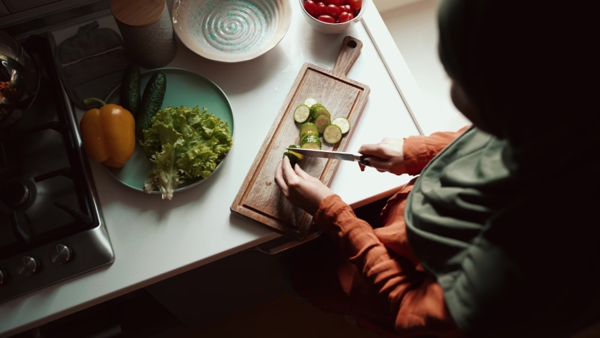 Overhead view of Muslim woman wearing hijab cuts cucumber in the kitchen