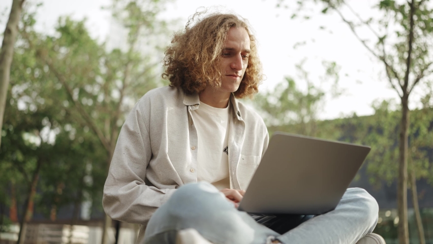 Positive curly-haired man working on laptop while sitting outdoors