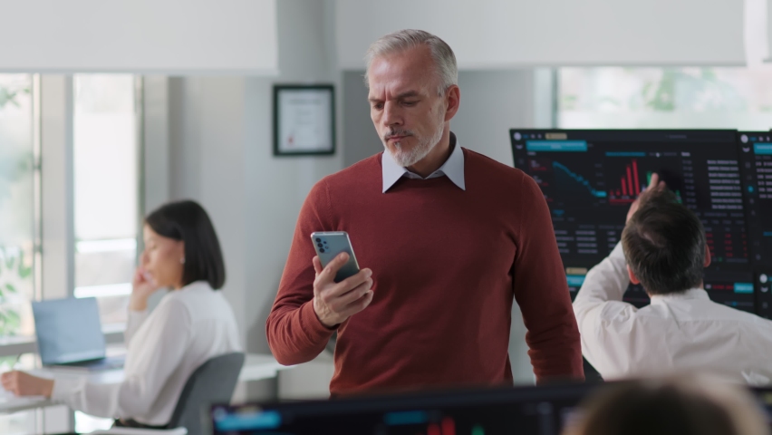 Mature gray haired man entrepreneur look at cellphone screen feeling stressed standing in modern office. Upset banker casual wear check stock market data on smartphone  | Shutterstock HD Video #1091221435