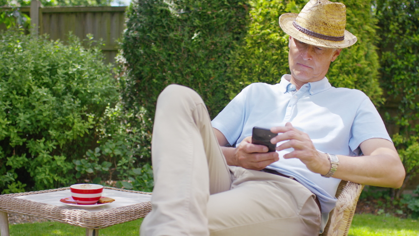 Handsome retired white male sips from a cup in his garden as he smiles at something he sees on his cell phone, in slow motion Royalty-Free Stock Footage #1091222289