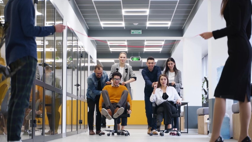 Cheerful people have fun during workday break. Two teams compete in chair on wheels racing in office hallway. Colleagues pushing coworkers on chairs. Winners rejoice. Employees film on smartphones. Royalty-Free Stock Footage #1091223257