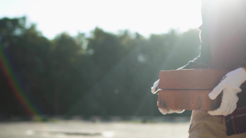 The builder girl carries bricks blocks of bright orange color, the hands of the builder in gloves carry the load of the warehouse of red bricks, a pile of bricks, the builder in a red bright jacket
