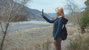 A slender and beautiful female tourist with a hiking backpack takes videos of mountains and rivers on her phone. The girl is resting in nature and admires the fascinating landscape.