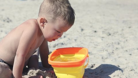 little baby boy child running towards sea ocean sand, playing on beach filling bucket with seawater, standing in water. happy family summer vacation concept. caucasian toddler kid on beach sunny day