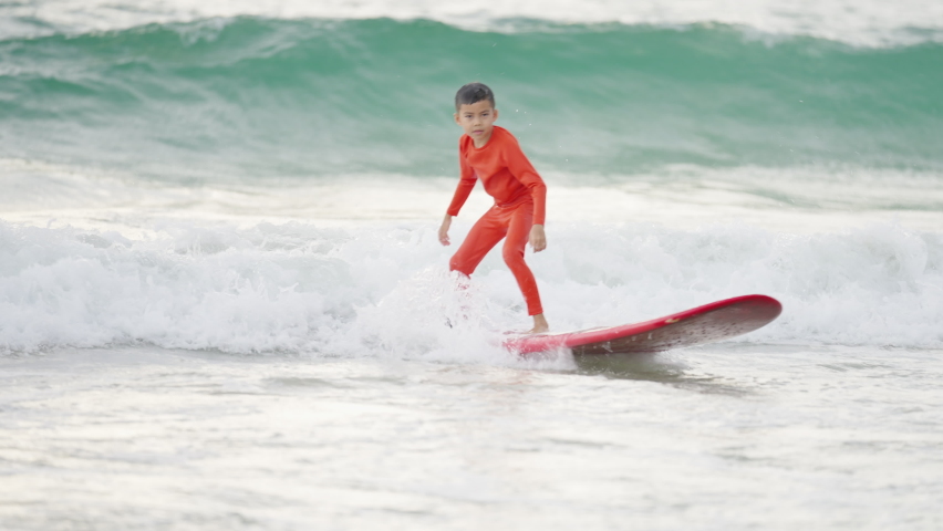 Little Asian boy surfer paddling surfboard and riding the wave in the sea on tropical beach at summer sunset. Children kid enjoy outdoor activity lifestyle and water sport surfing on summer vacation