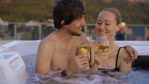 A young man and woman are relaxing in the hot tub on a rooftop with a view on mountains during sunset
