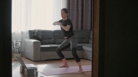 Side view of young woman wearing sportswear doing exercises and watching online tutorials on laptop while training on yoga mat in living room at home
