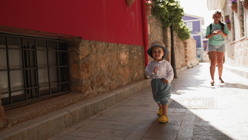 Toddler in hat walks with mom on narrow street with old vintage buildings. Cute little boy enjoys travel in touristic town on sunny summer day slow motion
