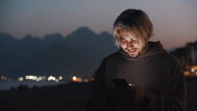 Blonde man in beige hoodie texting on his phone and looking around on the night beach - Powered by Shutterstock - Get 15% off with code: PIKWIZARD15