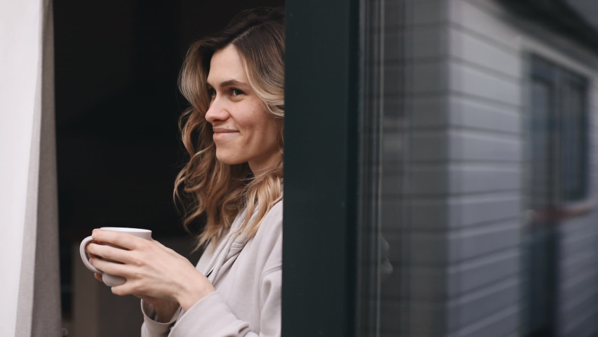 Smiling young woman waving with a friendly cheerful smile to her, Girl standing near glass door, holding mug with coffee or tea in her house and waving her hand. Curly woman  nice to meet her friends. Royalty-Free Stock Footage #1091251817