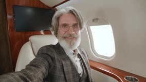 Happy mature millionaire man talking to camera holding phone recording vlog while flying on private jet. Gray-haired stylish senior man video calling in online chat using smartphone at airplane.