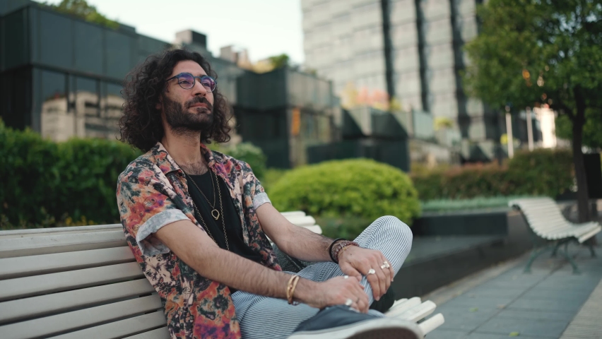 Relaxed curly-haired bearded man in eyeglasses looking around while sitting on the bench outdoors Royalty-Free Stock Footage #1091261291