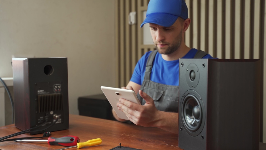 Master in overalls diagnoses music column checking information in tablet sitting at wooden desk. Repair of electronics at home. Broken device on table