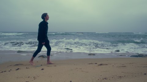 Barefoot  young man walks on the empty ocean beach on the summer cloudy rainy weather. Sad guy walks along a sandy beach at high tide. Human feels loneliness and melancholy, outdoors. 4k cinematic