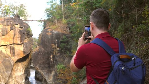 Casual travel blogger shooting video of amazing nature in national park in Thailand. Back view of 30s forest hiker photographer man with backpack make video of canyon for social media on mobile phone