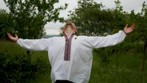 Adult caucasian male wearing Ukrainian traditional shirt spreading hands to sides and looking up outdoors. Man in embroidered vyshyvanka looking to the sky and talking to God with open arms in nature
