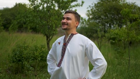 Handsome caucasian man wearing a beautiful white vyshyvanka outdoors. Cheerful young slav dude in traditional embroidered Ukrainian and Belarussian shirt smiling and enjoying nature in grassy meadow

