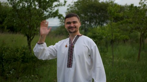 Smiling caucasian man in traditional Eastern European embroidered vyshyvanka turning around and waving hand to viewer. Adult white male in authentic long shirt friendly greeting and waving hello
