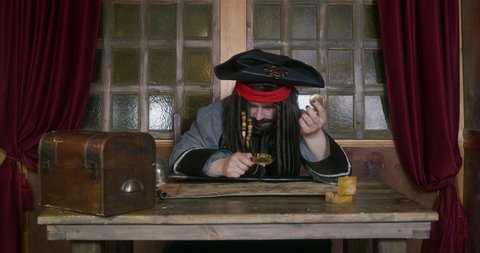 Pirate in tricorn hat and wig with dreadlocks decorated with beads studies map through magnifying glass, searching of place where treasure was buried, checking compass. He nods approvingly