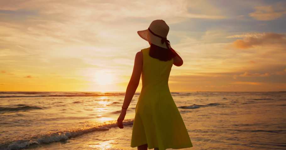 Slow motion asian woman in beautiful yellow dress walking by beach at golden sunset - Female tourist on summer vacation