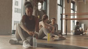 Slowmo of young Black woman in activewear drinking water while sitting on mat in fitness studio, resting after training. Two girls chatting in background - Powered by Shutterstock - Get 15% off with code: PIKWIZARD15