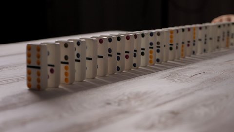 White dominoes with multicolored dots slowly fall on a light wooden background. The background is blurred. Concept of domino effect and chain reaction.