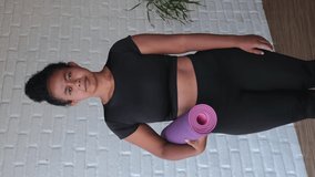 A dark-skinned woman in a rolled-up lilac yoga mat stands standing, smiling sweetly. Her smile is impressive. Metis woman is ready to practice yoga. Zambo model. vertical video.