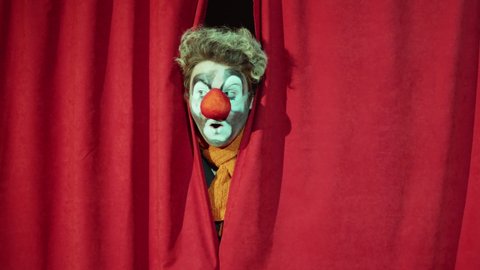 Clown peeking through red stage curtains and performing comedy in circus