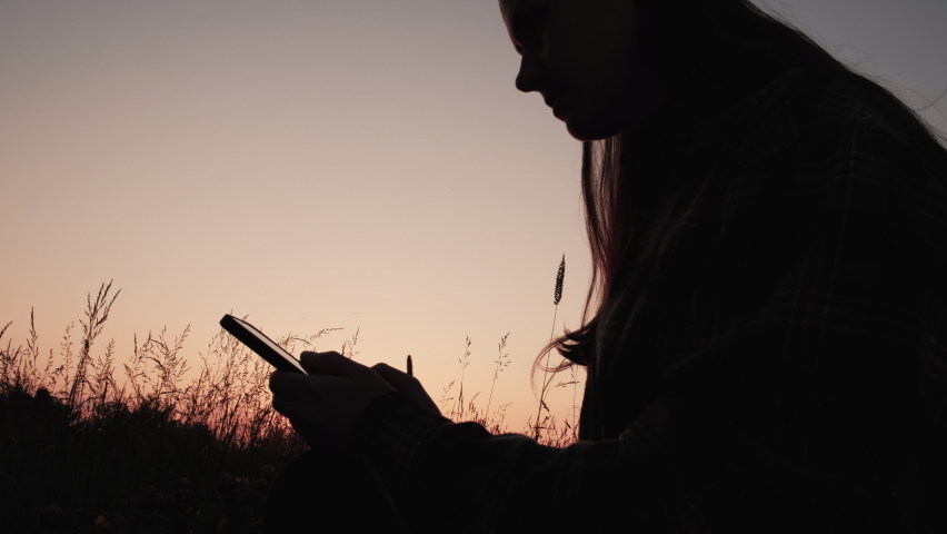 Silhouette of young woman sitting in grass and holding mobile phone during sunset in mountains, enjoying nature. Smartphone application market, podcast listening. Adventure, nature, relaxation concept