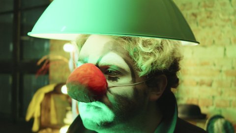 Tilt down shot of clown with makeup and red nose dancing with hanging lamp on his head in dressing room