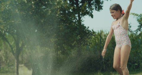 Cinematic authentic shot of carefree happy little girl is having fun to run, jump and splash herself with garden water sprinkler on green nature background in hot sunny summer day.