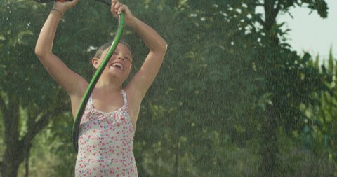 Cinematic authentic close up shot of carefree happy little girl is having fun to run, jump and splash herself with garden water sprinkler on green nature background in hot sunny summer day.