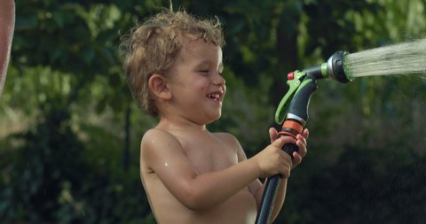 Cinematic authentic close up shot of carefree happy toddler boy and his sisters are having fun to run, jump and splash with garden water sprinkler on green nature background in hot sunny summer day.