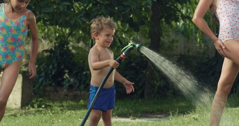 Cinematic authentic shot of carefree happy toddler boy and his sisters are having fun to run, jump and splash with garden water sprinkler on green nature background in hot sunny summer day.