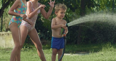 Cinematic authentic shot of carefree happy toddler boy and his sisters are having fun to run, jump and splash with garden water sprinkler on green nature background in hot sunny summer day.