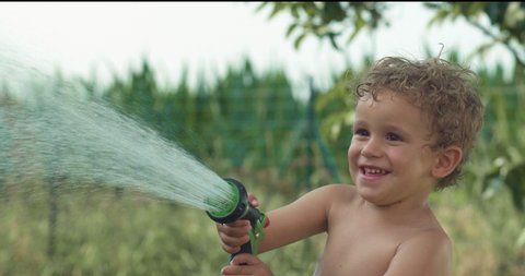 Cinematic authentic close up shot of carefree happy smiling toddler boy  having fun to play with garden water sprinkler on green nature background in hot sunny summer day.