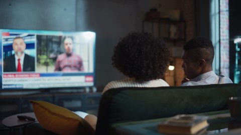 Black Couple Watches Evening News on TV and Sharing Their Opinions while Sitting on Couch at Home in the Evening. Spouses Spend Time Together. Back View Shot