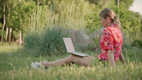 A middle-aged woman sits on the grass in the park. Using a video link on a laptop, she conducts a conversation. High quality 4k footage