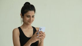 A Asian young fitness woman holding smartphone relaxing after workout at home. Smiling Indian female using cell phone video calling while exercise.