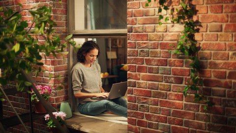 Creative Female Writer Works on a Laptop Computer while Sitting on a Windowsill of her Cozy Apartment. Professional Woman Artist Working Remotely from Home, Listens Birds Sing. Outside Building Window