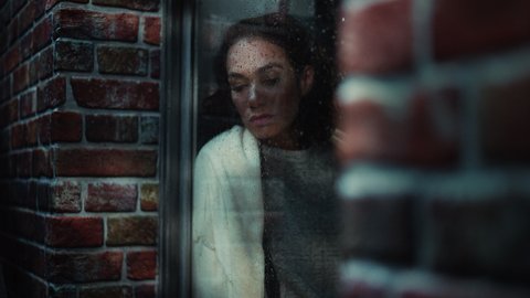 Sad Woman in Melancholic Mood and Feeling Depressed. Hopeless Latin Female Sitting on a Windowsill, Looking outside Window on a Rainy Street. Loneliness Concept. Inside Apartment Window Shot