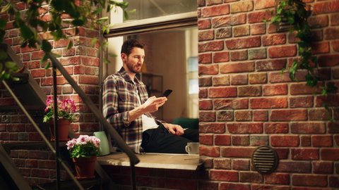 Happy Handsome Caucasian Man Using Smartphone while Sitting on a Windowsill of his Cozy Apartment. Casual Shirt Wearing Guy Browsing Internet, Checking Videos on Social Media, Having Fun on Sunny Day