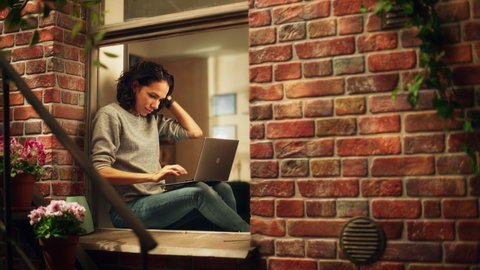 Creative Female Works on a Laptop Computer while Sitting on a Windowsill of her Cozy Apartment. Professional Woman Working Remotely from Home, Listens Birds Sing. Outside Building Window Shot