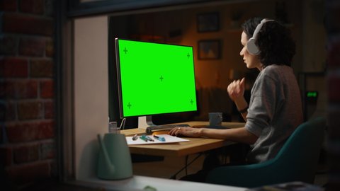 Female Designer Using Desktop Computer with Green Screen Chroma Key Monitor. Remote Access Manager Listening to Music or Podcast in Headphones, Freelancer Working at Night. Zoom Out Window Shot