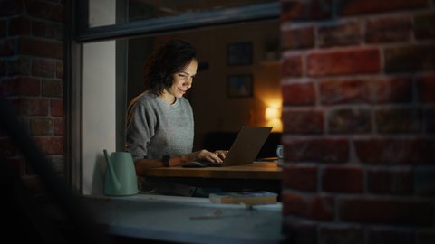 Smiling Hispanic Woman at Home Using Laptop on the Warm Cozy Evening. Remote Work Professional Freelancer Working From Home. Shot Inside Apartment Window