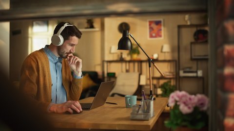 Thoguhtful Professional Man Using Laptop, Listens Educational Audio Podcast through Headphones while Working Remotely in Home office. Stylish Professional Remote Access Work. Inside Apartment Shot