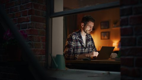 Focused Man Working on a Laptop, late in the Evening Finishing Important Project in his Home Office. Freelancer Working Remotely. Warm Lit Cozy Apartment with Inside Window Static Shot
