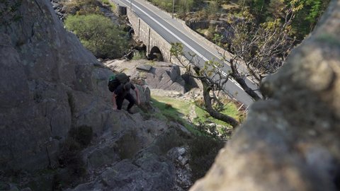 A photographer and backpacker climbing a steep rocky mountainside with a road in the background