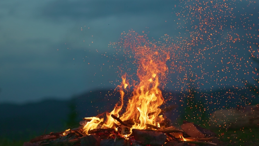 Campfire at dusk against the backdrop of mountains and forest. Tourist bonfire with sparks flying in the air, slow motion shot. Royalty-Free Stock Footage #1091293587